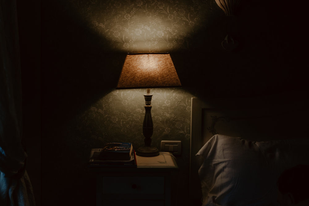 Table lamp on small bed-side table, casting light from above and below its shade onto a patterened-wallpaper wall behind it, in an otherwise dark room with a bed - of which only the headboard and a striped pillow can be seen.